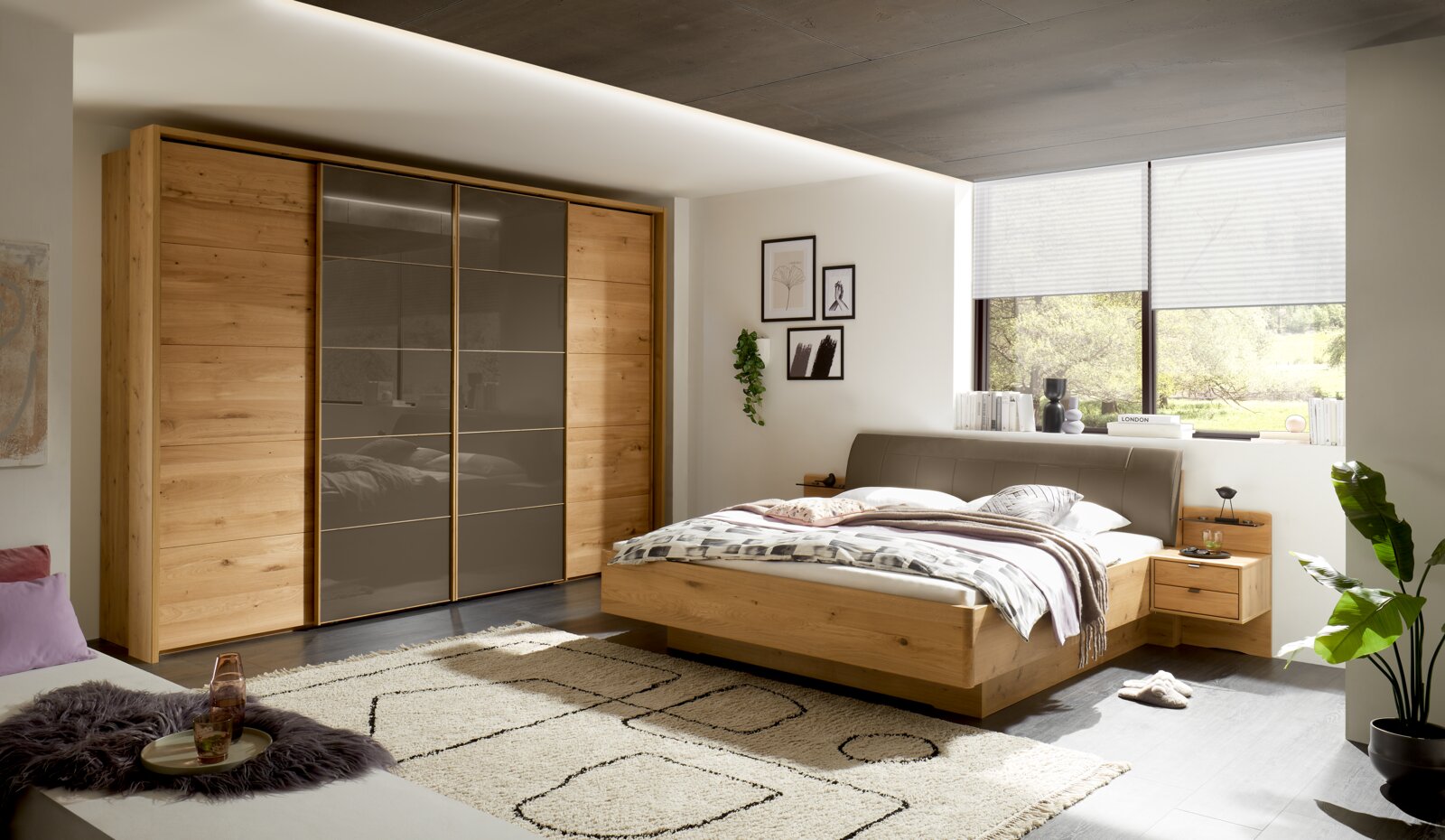 Linea Natura Schwebetürenschrank 4-türig CARDIFF Moderner Schwebetürenschrank mit vier Türen in Holzoptik und grauen Glaselementen, seitliche Perspektive im Schlafzimmer.