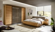 Moderner Schwebetürenschrank mit vier Türen in Holzoptik und grauen Glaselementen, seitliche Perspektive im Schlafzimmer.