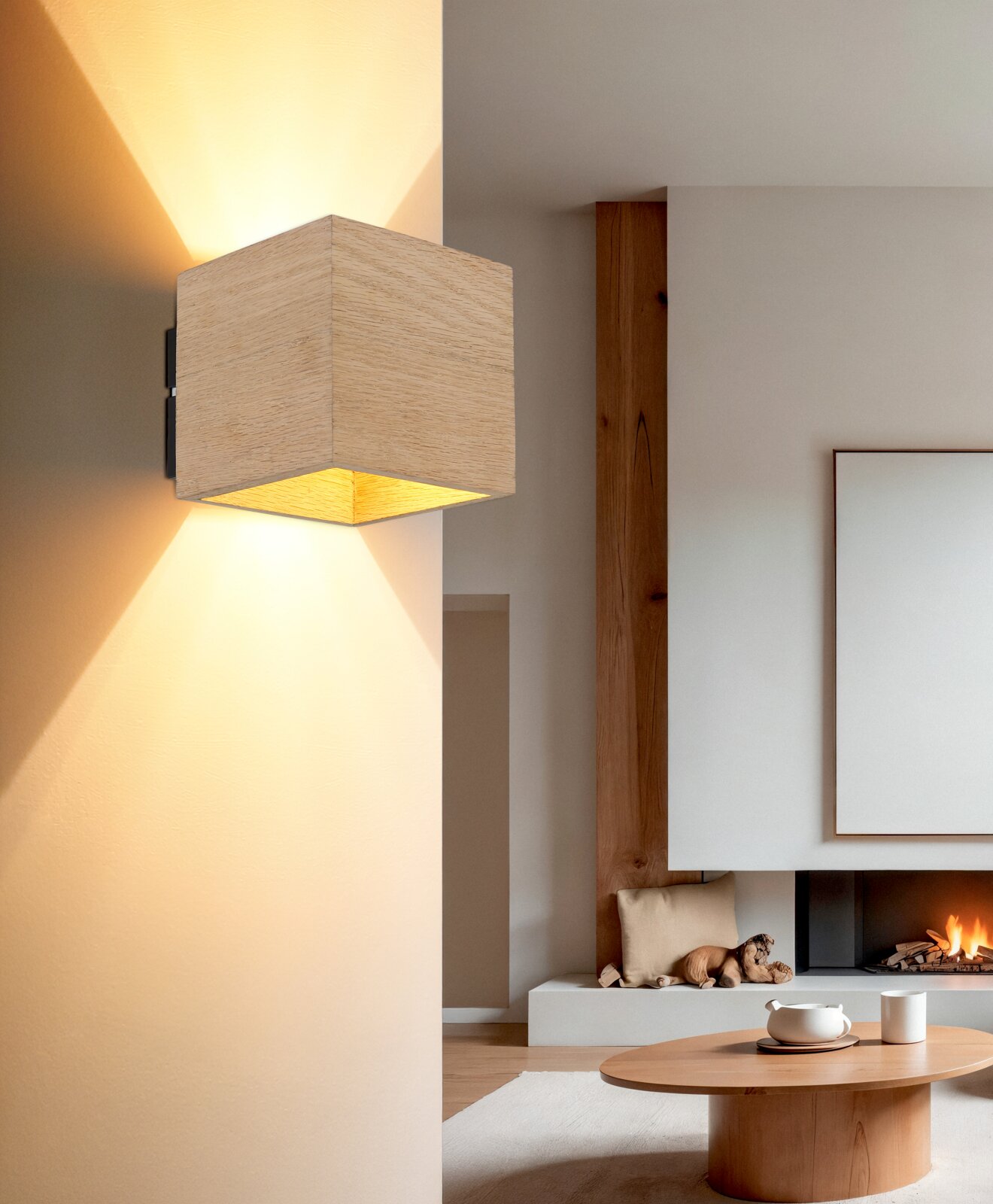NOWA Wandlamp EDWIN Kubusvormige wandlamp van licht hout met warm licht naar boven en naar beneden, bevestigd aan een witte muur; zijdelingse close-up vanuit een licht schuin perspectief; op de achtergrond een moderne woonkamer met open haard.
