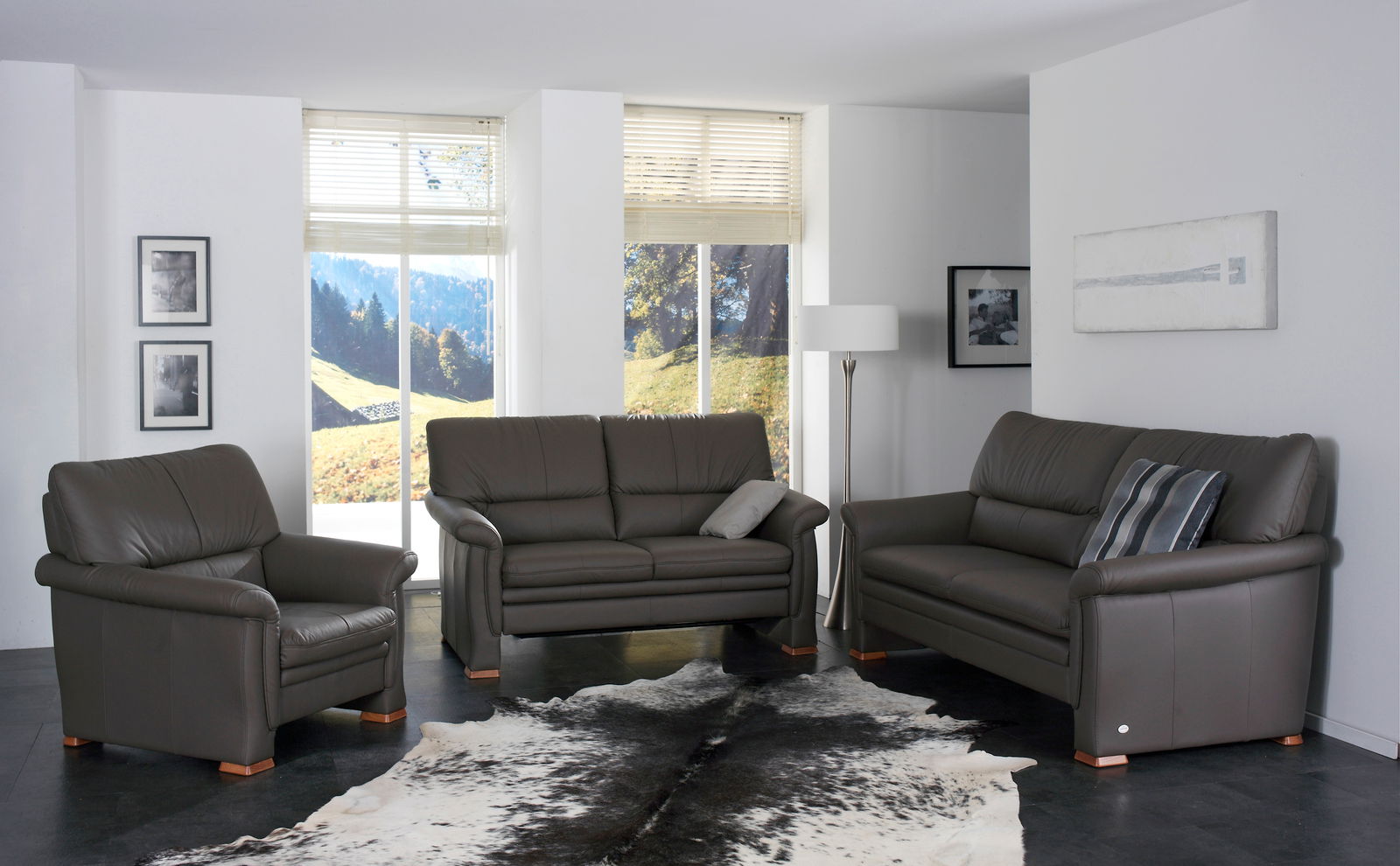 Wohnzimmer mit einem 2,5-sitzigen Himolla Polster 2253 DAYTON SLEEPOLY Sofa in braunem Leder, frontal betrachtet, umgeben von einem passenden Sessel und einem weiteren Sofa, mit Blick auf eine Landschaft durch große Fenster.