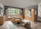 Ankleidebank aus Holz mit gepolsterter Sitzfläche, platziert vor einem Bett in einem modernen Schlafzimmer mit großen Fenstern. Perspektive von vorne.