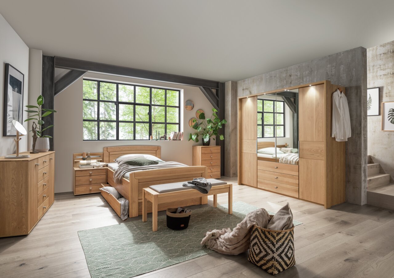 Ankleidebank aus Holz mit gepolsterter Sitzfläche, platziert vor einem Bett in einem modernen Schlafzimmer mit großen Fenstern. Perspektive von vorne.