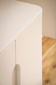 Detailaufnahme einer weißen, minimalistischen Schrank- oder Sideboard-Ecke mit abgerundeter Kante und eingelassener Griffmulde, fotografiert aus leicht erhöhter schräger Vorderansicht vor Holzboden.