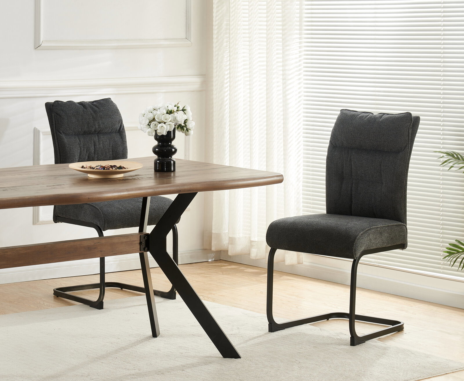MID.YOU Schwingstuhl 2er-Set FRIEDA Moderner Essbereich mit Holztisch und zwei dunkelgrauen Polsterstühlen mit schwarzem Freischwinger-Gestell, seitliche Perspektive auf Tisch und Stühle, dekoriert mit Schale und Blumen auf hellem Teppich vor Fensterjalousien.