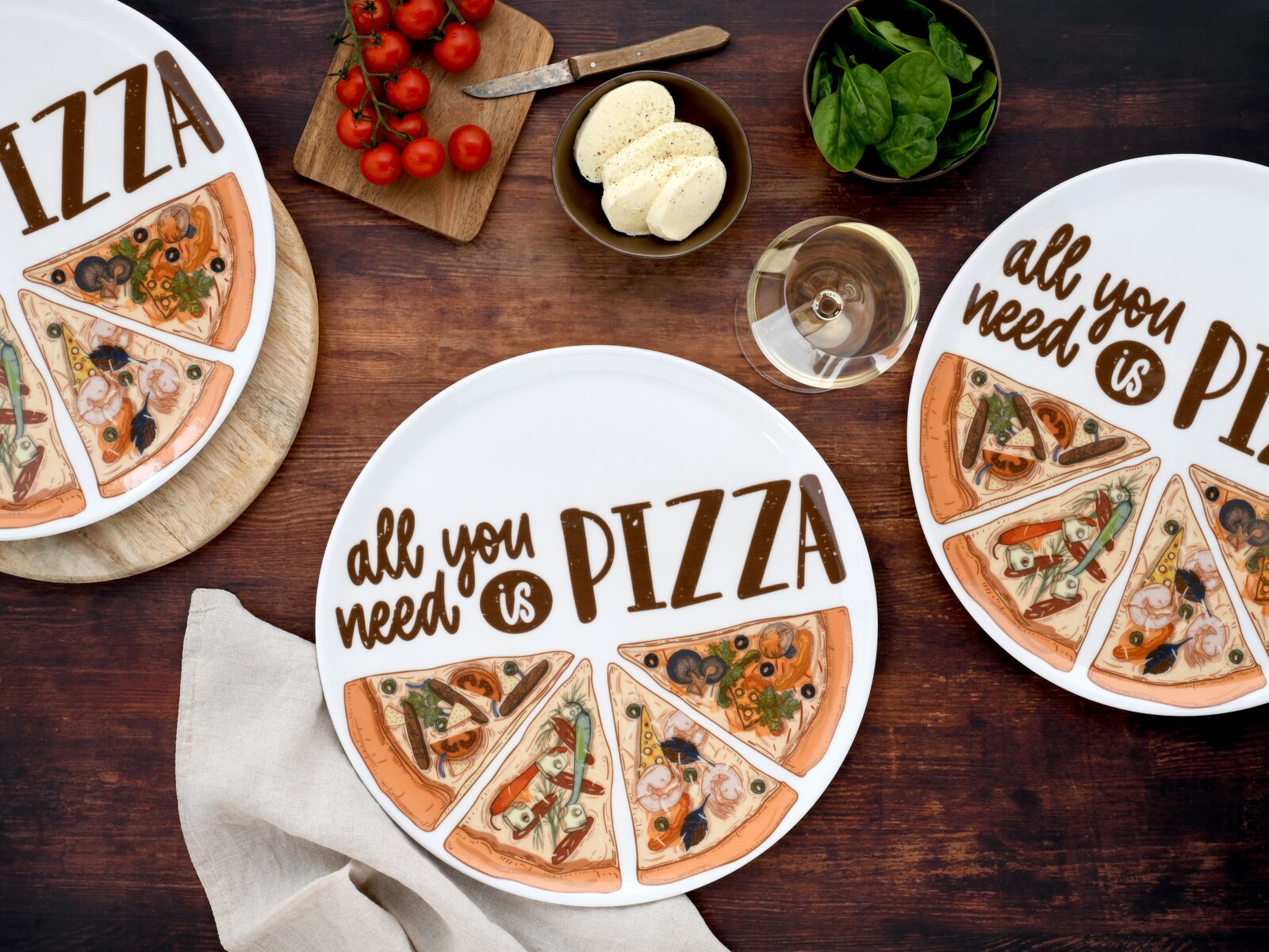 Draufsicht auf einen runden weißen Pizza-Teller mit der Aufschrift „all you need is PIZZA“ und illustrierten Pizzastücken; daneben auf Holztisch Cherrytomaten, Mozzarella, Spinat und ein Glas Weißwein.