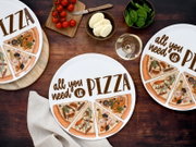 Draufsicht auf einen runden weißen Pizza-Teller mit der Aufschrift „all you need is PIZZA“ und illustrierten Pizzastücken; daneben auf Holztisch Cherrytomaten, Mozzarella, Spinat und ein Glas Weißwein.