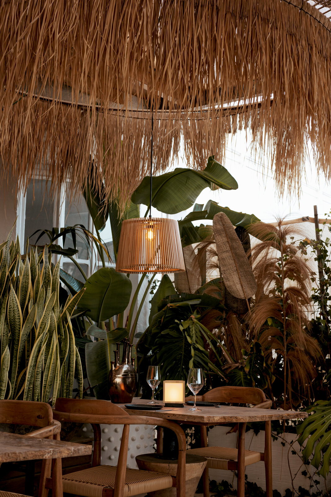Gezellig restaurantgedeelte met een hangende rotan-pendellamp onder een rieten dak, een houten tafel met een kaars en wijnglazen, omringd door weelderige tropische planten; perspectief: licht naar boven gerichte binnenopname met uitzicht op het plafond en
