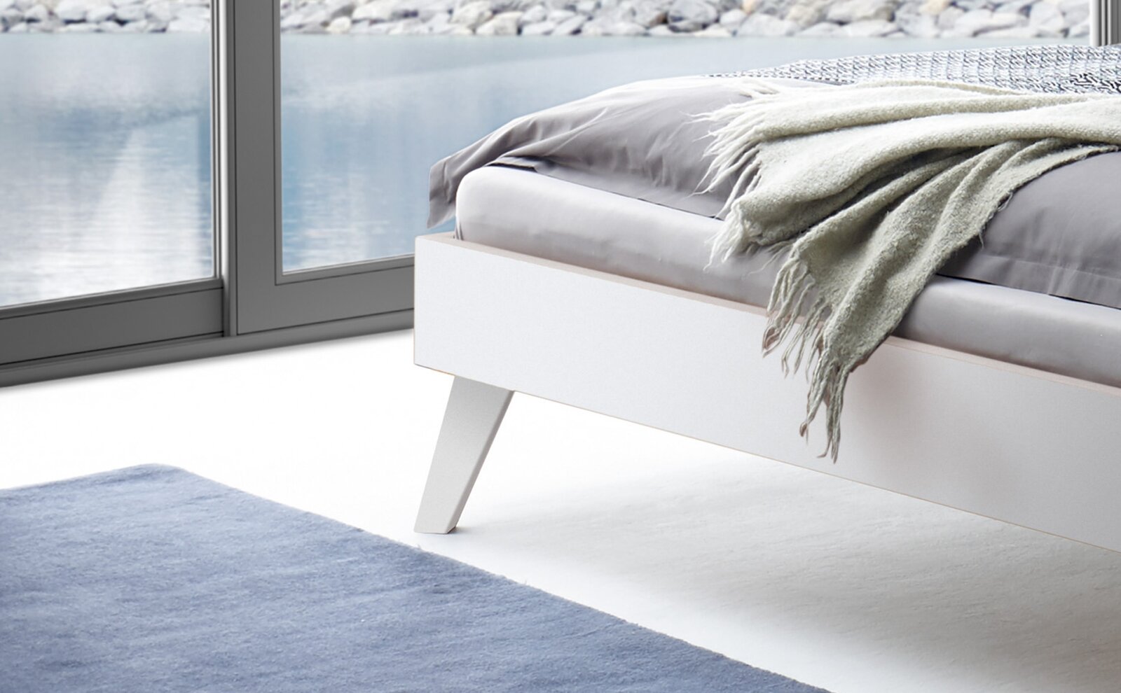 Modernes weißes Bettgestell in einem hellen Raum mit Blick auf ein Fenster, das auf einen See zeigt. Perspektive von der Seite.