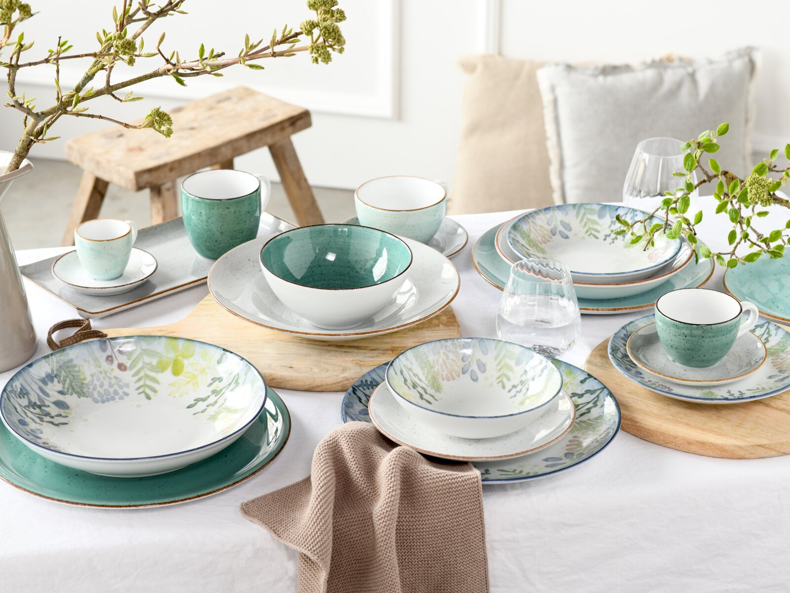 Schräg von vorn fotografiertes, gedecktes Tisch-Arrangement mit Keramik-Geschirrset in Weiß und Türkis/Grün, teils mit floralen Mustern, inklusive Tassen, Schalen und Tellern sowie Gläsern auf weißer Tischdecke.