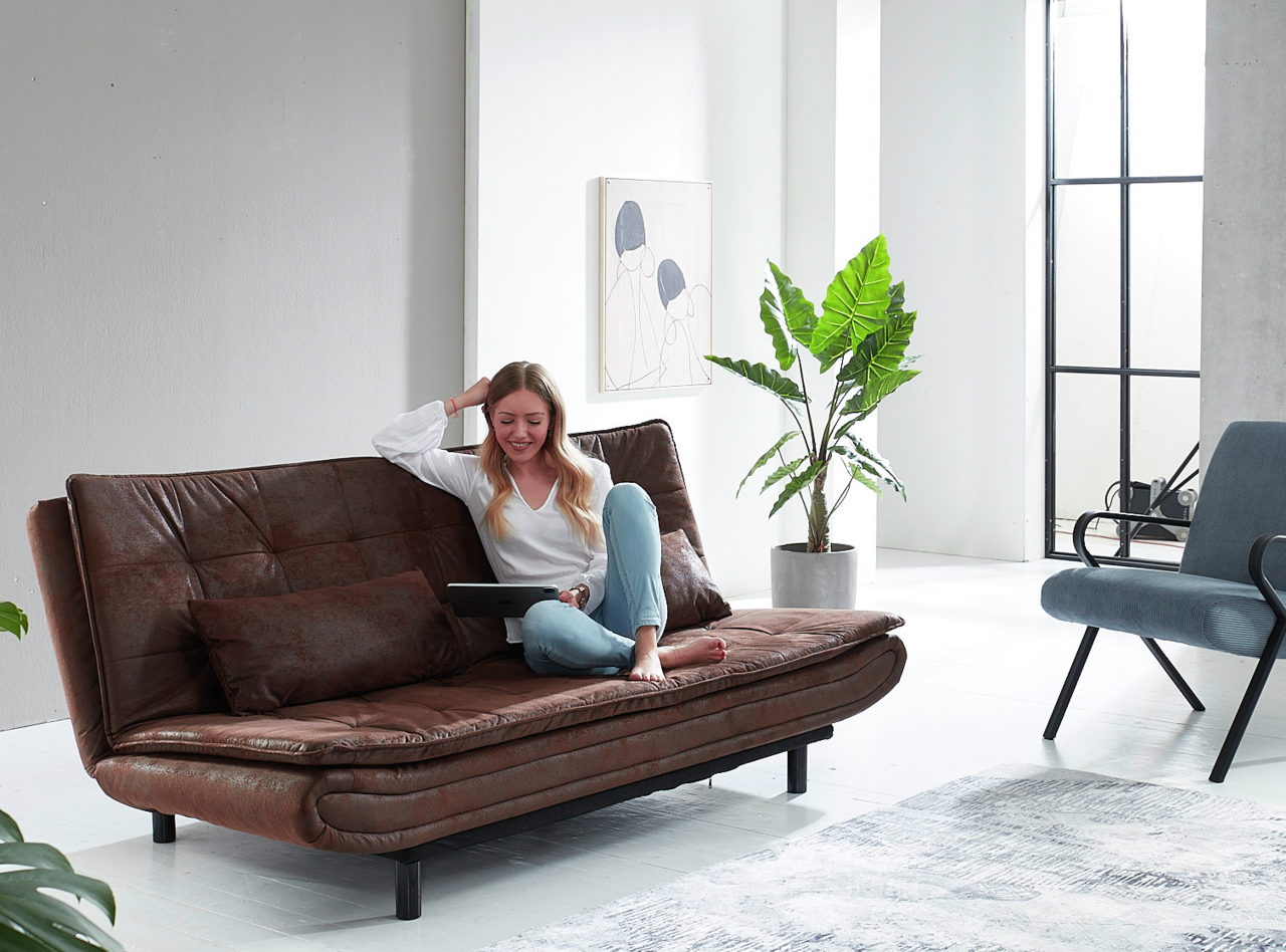 Eine Frau sitzt entspannt auf einem braunen Sessel im modernen Wohnzimmer, seitliche Perspektive.