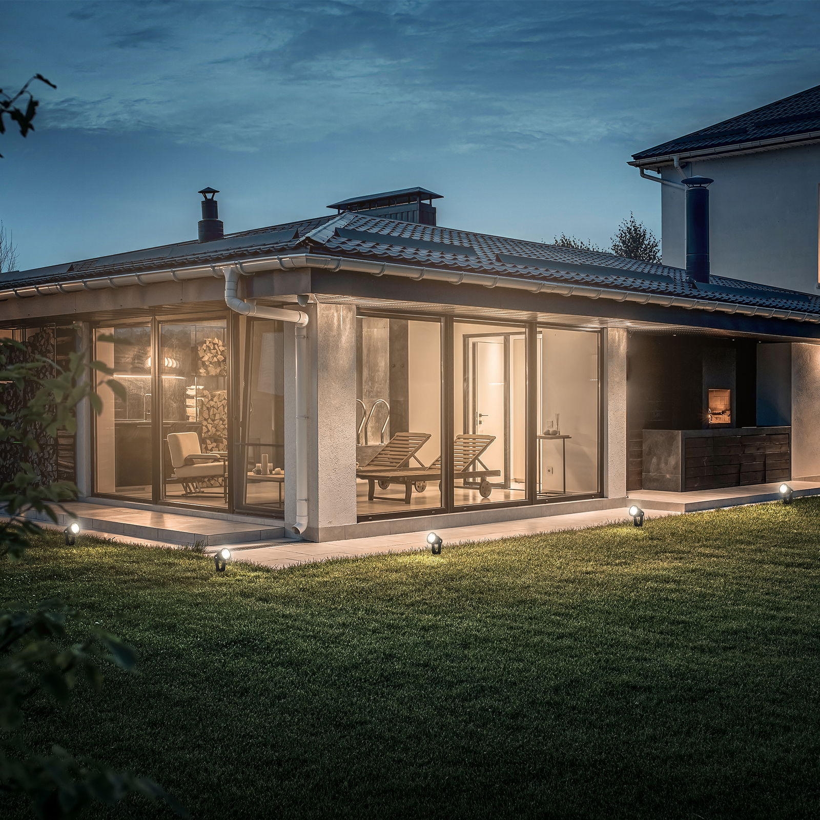 Gartenbeleuchtung mit Erdspieß-Spot-Verlängerungsset, seitliche Perspektive auf ein modernes Haus mit großen Glasfenstern und beleuchtetem Garten.