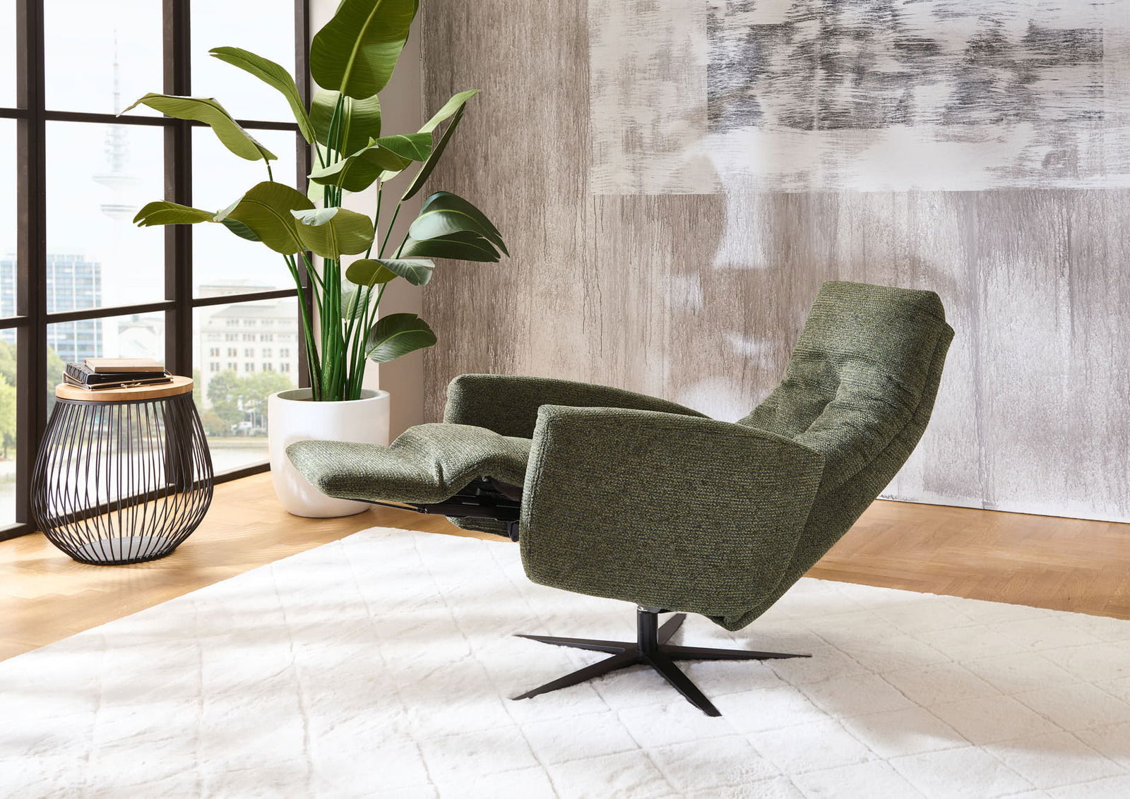 Grüner Relax-Sessel in seitlicher Perspektive, neben einem Beistelltisch und einer großen Zimmerpflanze, in einem modernen Wohnzimmer mit großen Fenstern.