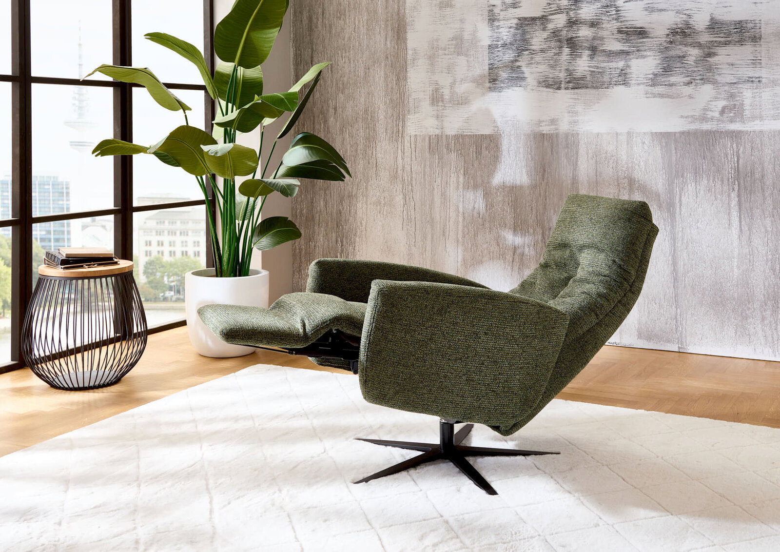 Grüner Relax-Sessel in seitlicher Perspektive, neben einem Beistelltisch und einer großen Zimmerpflanze, in einem modernen Wohnzimmer mit großen Fenstern.