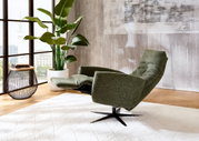 Grüner Relax-Sessel in seitlicher Perspektive, neben einem Beistelltisch und einer großen Zimmerpflanze, in einem modernen Wohnzimmer mit großen Fenstern.