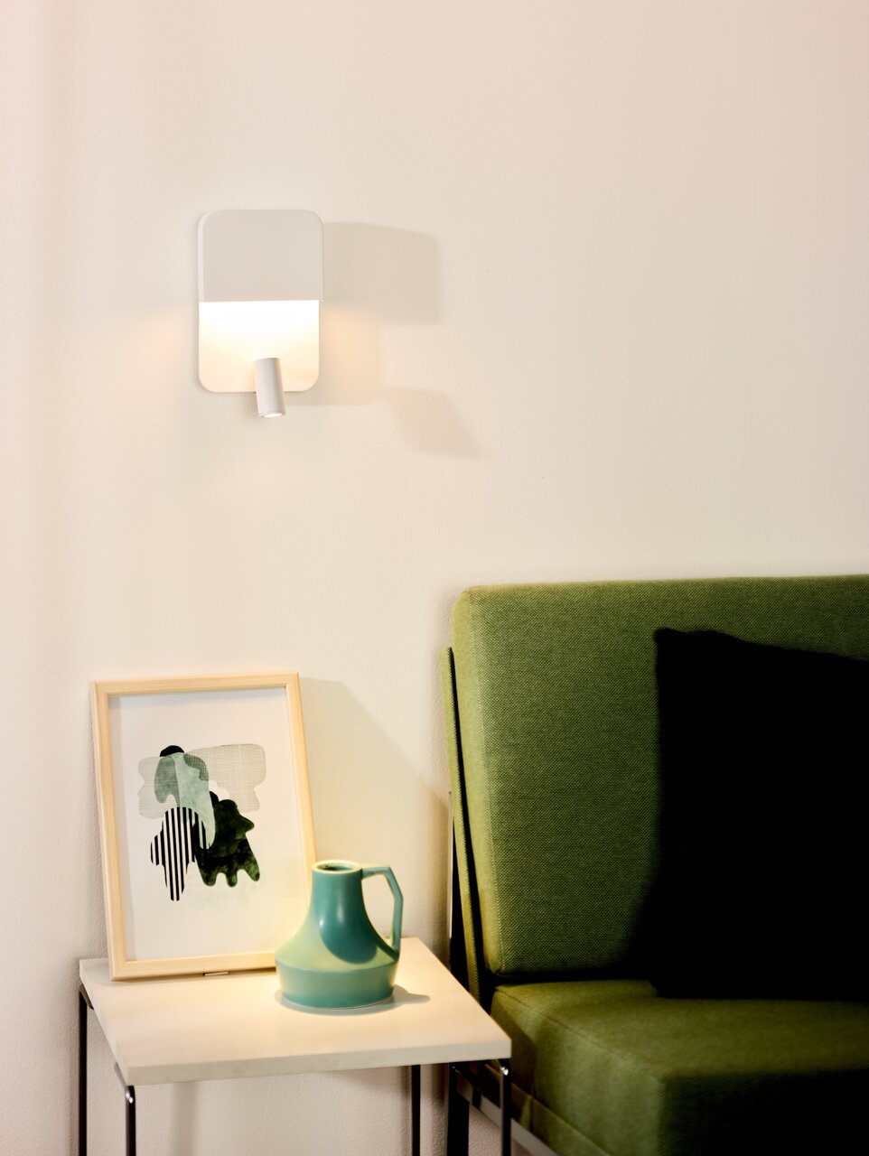 Lucide Wand-/Bettleuchte BOXER Moderne Wand-Bettleuchte in minimalistischem Design, montiert über einem Beistelltisch mit Kunstwerk und Vase, neben einem grünen Sofa. Perspektive von vorne.
