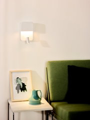 Moderne wandlamp in minimalistisch design, bevestigd boven een bijzettafeltje met een kunstwerk en een vaas, naast een groene bank. Aanzicht van voren.