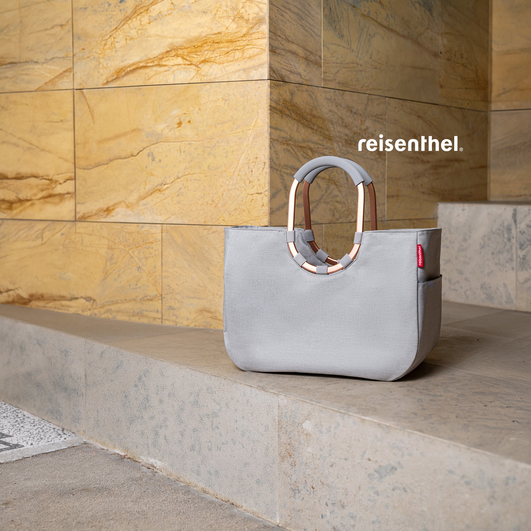 Graue Reisenthel-Tasche mit runden Griffen auf Steinbank vor moderner Natursteinwand.