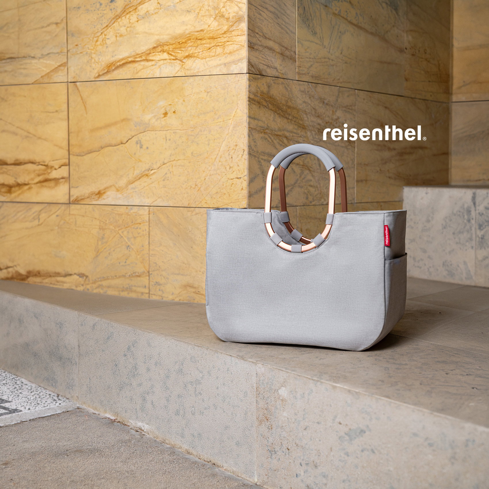 Graue Reisenthel-Tasche mit runden Griffen auf Steinbank vor moderner Natursteinwand.