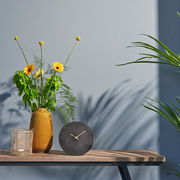 Moderne schwarze Tischuhr mit goldenen Zeigern, frontal auf einem Holztisch neben einer gelben Vase mit Blumen und einem Korb platziert.