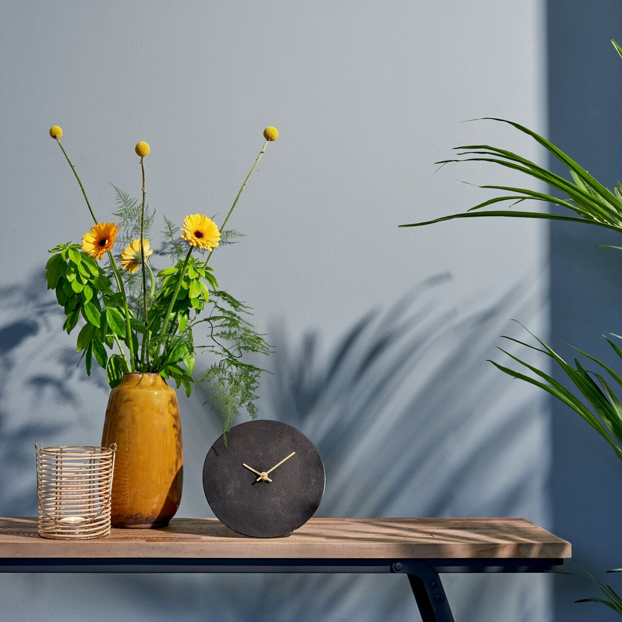 Moderne zwarte tafelklok met gouden wijzers, vooraan op een houten tafel geplaatst naast een gele vaas met bloemen en een mand.
