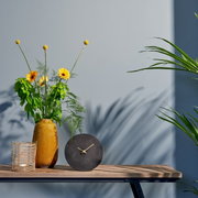 Moderne zwarte tafelklok met gouden wijzers, vooraan op een houten tafel geplaatst naast een gele vaas met bloemen en een mand.