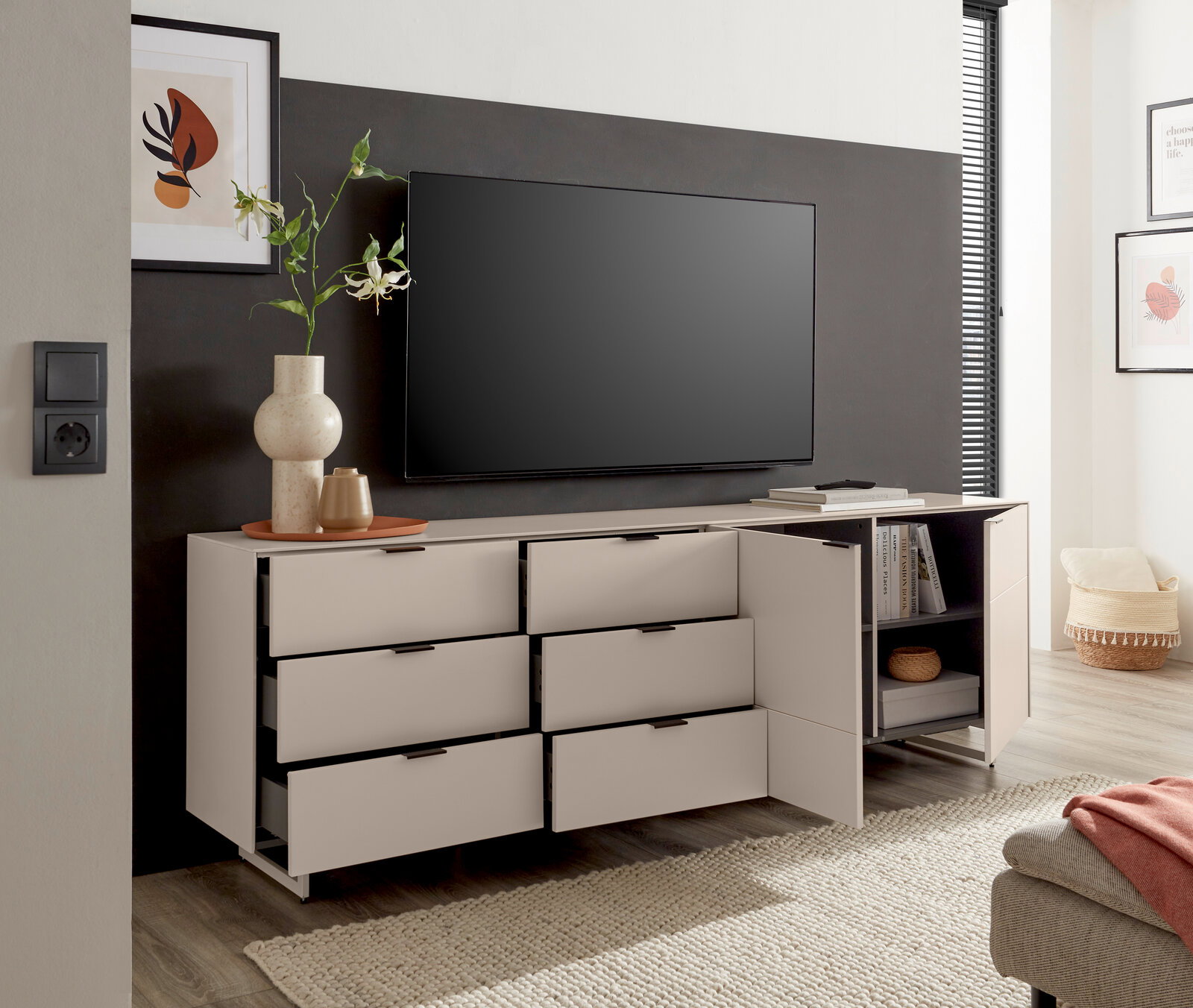 Modernes weißes TV-Lowboard/Sideboard mit sechs Schubladen und offenem Regal, eine Tür und mehrere Schubladen geöffnet, mit Fernseher darüber; Perspektive: frontale Ansicht leicht von links.