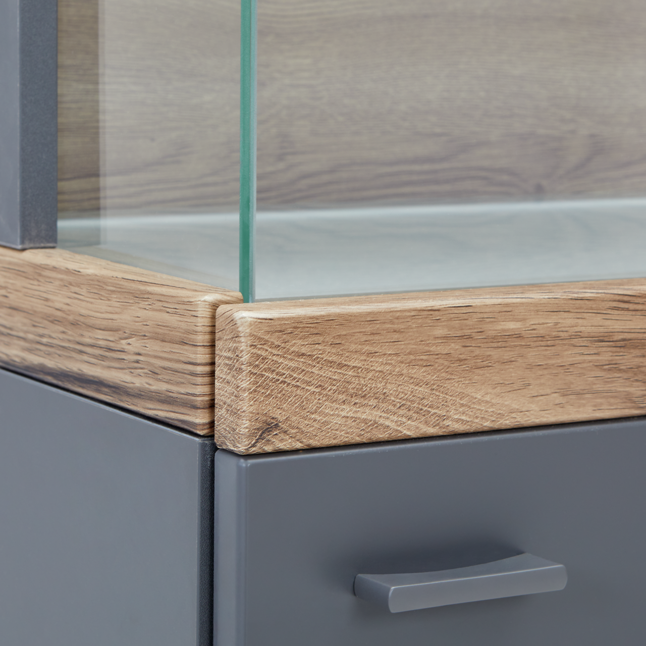 Nahaufnahme einer Vitrine mit Glasausschnitt, Holzrahmen und grauer Schublade, Perspektive von der Ecke.