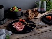 Schwarzes, mattes Geschirr-Set mit Teller und Schalen auf rustikalem Holztisch, angerichtet mit Prosciutto, Cherrytomaten und Salat, daneben Rotweinglas und Brotbrett; Perspektive: leicht erhöhte seitliche Ansicht