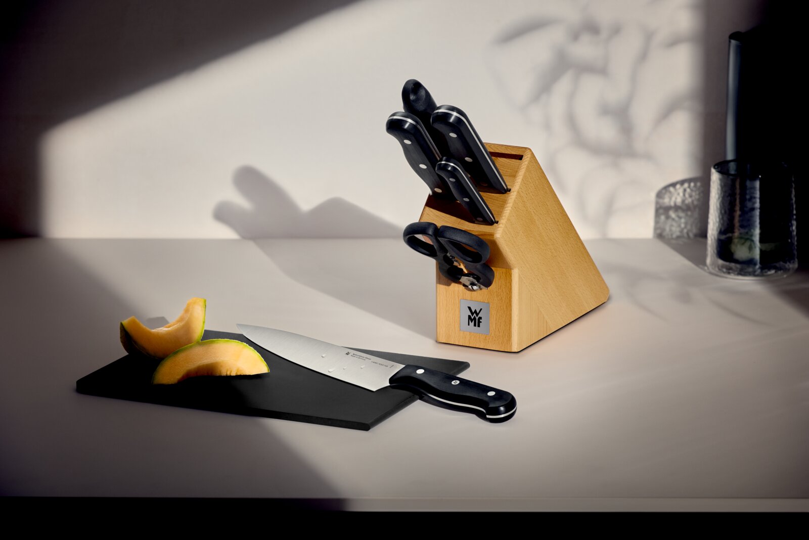WMF Messerblock 7-tlg. MEISTERKLASSE WMF Messerblock aus hellem Holz mit mehreren Messern und Küchenschere; vorne liegt ein Kochmesser neben Melonenstücken auf einem schwarzen Schneidebrett, aufgenommen in seitlicher Perspektive