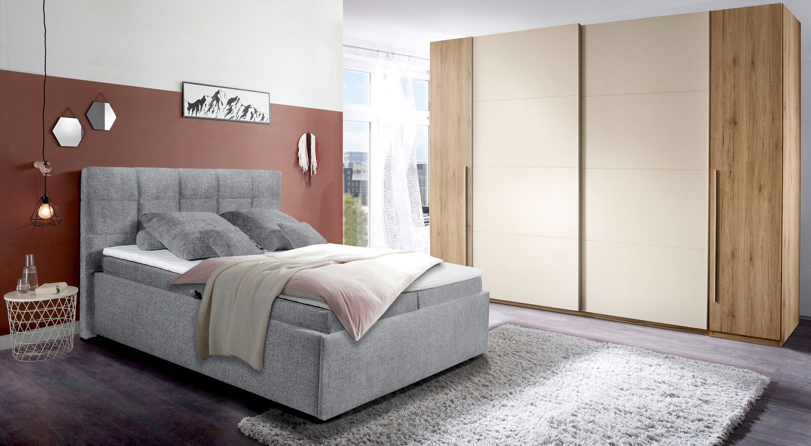 Graues Polsterbett mit hohem Kopfteil, dekoriert mit Kissen und Decke, in einem modernen Schlafzimmer mit Teppich und großem Kleiderschrank, seitliche Perspektive.