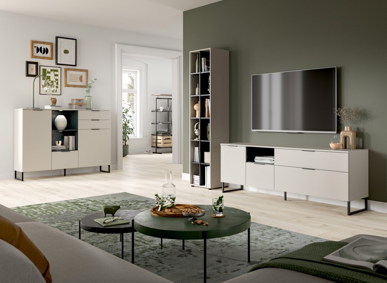 Modernes Wohnzimmer-Setup mit hellen Sideboards und TV-Lowboard auf schwarzen Metallfüßen, hohem Regal und runden Couchtischen auf grünem Teppich; Perspektive: Weitwinkelaufnahme aus leicht erhöhter, frontaler Raumecke.