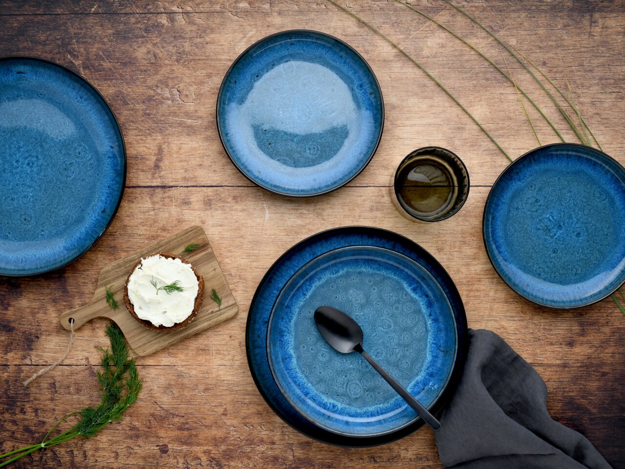 Vogelperspektive auf ein 18-teiliges Tellerset in Blau mit rustikalem Design, arrangiert auf einem Holztisch mit Besteck und Serviette.
