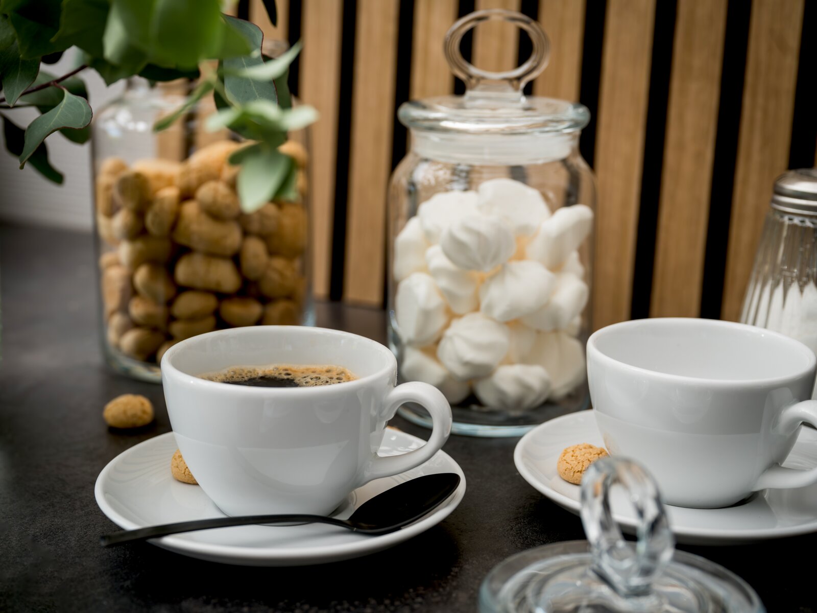 Schräg von vorn aufgenommene Szene mit zwei weißen Kaffeetassen auf Untertassen, eine mit schwarzem Kaffee, daneben Keks und Löffel; im Hintergrund Glasbehälter mit Baiser und Keksen sowie Zuckerdose.