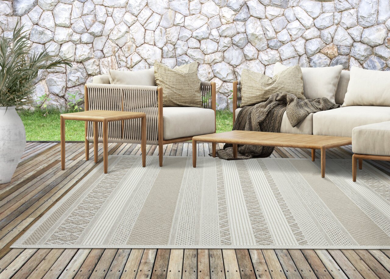 Moderner Outdoor-Teppich in Beige mit geometrischen Mustern auf einer Holzterrasse, umgeben von stilvollen Gartenmöbeln aus Holz und beigen Kissen, mit Blick auf eine Steinmauer im Hintergrund.