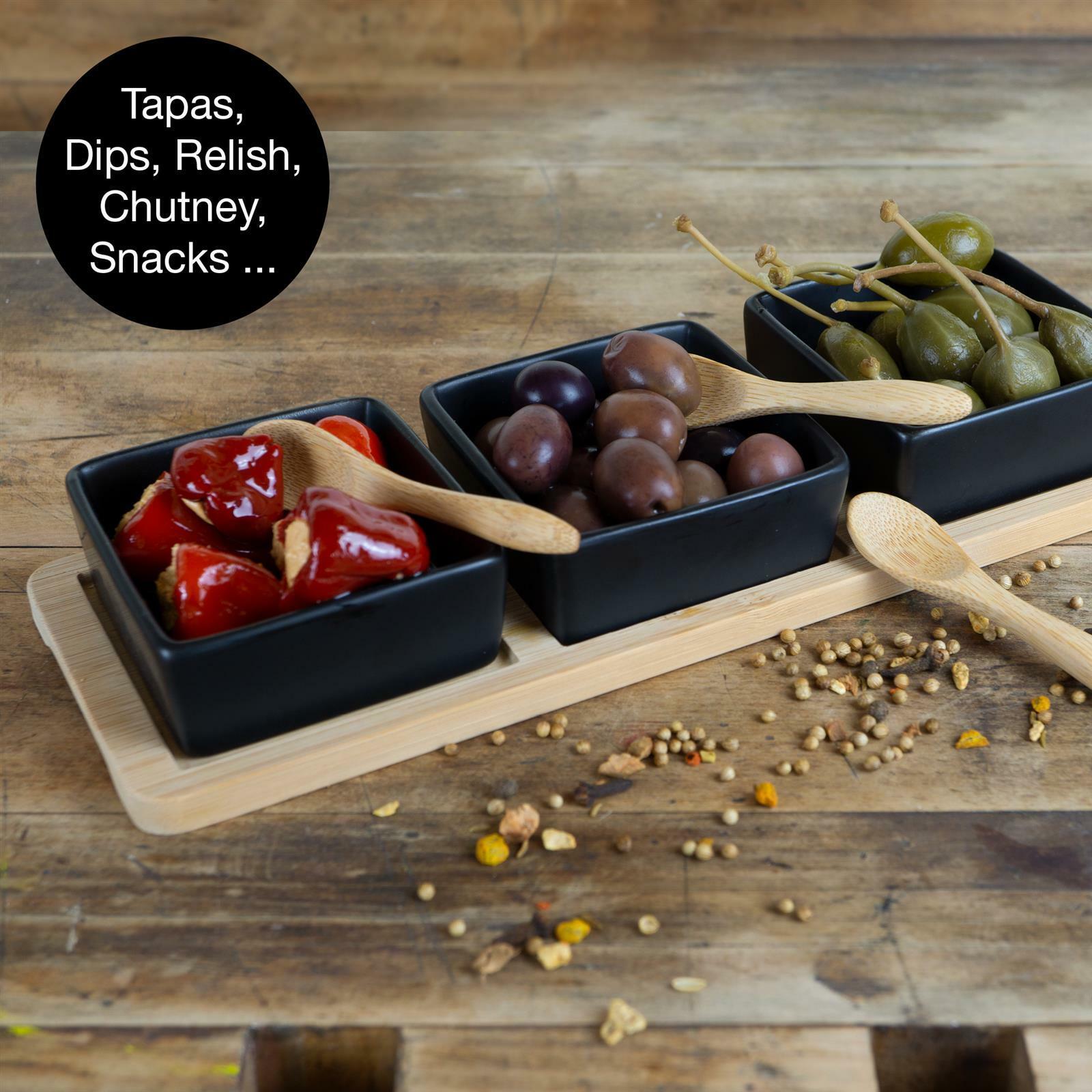 Servierset aus Bambus mit drei schwarzen Schalen, gefüllt mit gefüllten Paprikas, Oliven und Kapern, auf einem Holztisch von oben fotografiert.