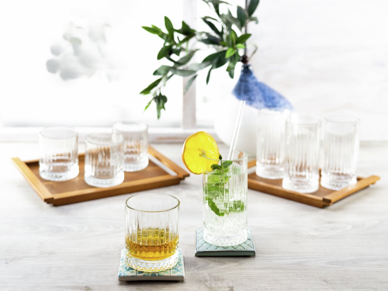 Ein Set aus Longdrinkbechern Elysia, präsentiert auf einem Tisch mit dekorativen Untersetzern und einem frischen Getränk mit Zitrone und Minze, aus einer seitlichen Perspektive fotografiert.