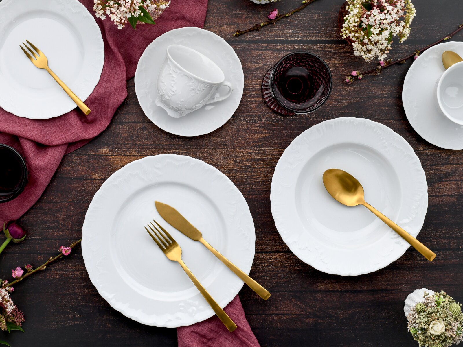 Draufsicht auf ein elegantes Tischgedeck mit weißen Porzellantellern und -tassen mit Reliefmuster, goldfarbenem Besteck und dunklen Gläsern auf rustikalem Holztisch mit bordeauxfarbenen Stoffservietten und Blumen.
