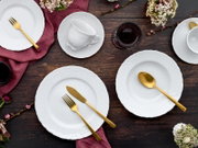 Draufsicht auf ein elegantes Tischgedeck mit weißen Porzellantellern und -tassen mit Reliefmuster, goldfarbenem Besteck und dunklen Gläsern auf rustikalem Holztisch mit bordeauxfarbenen Stoffservietten und Blumen.
