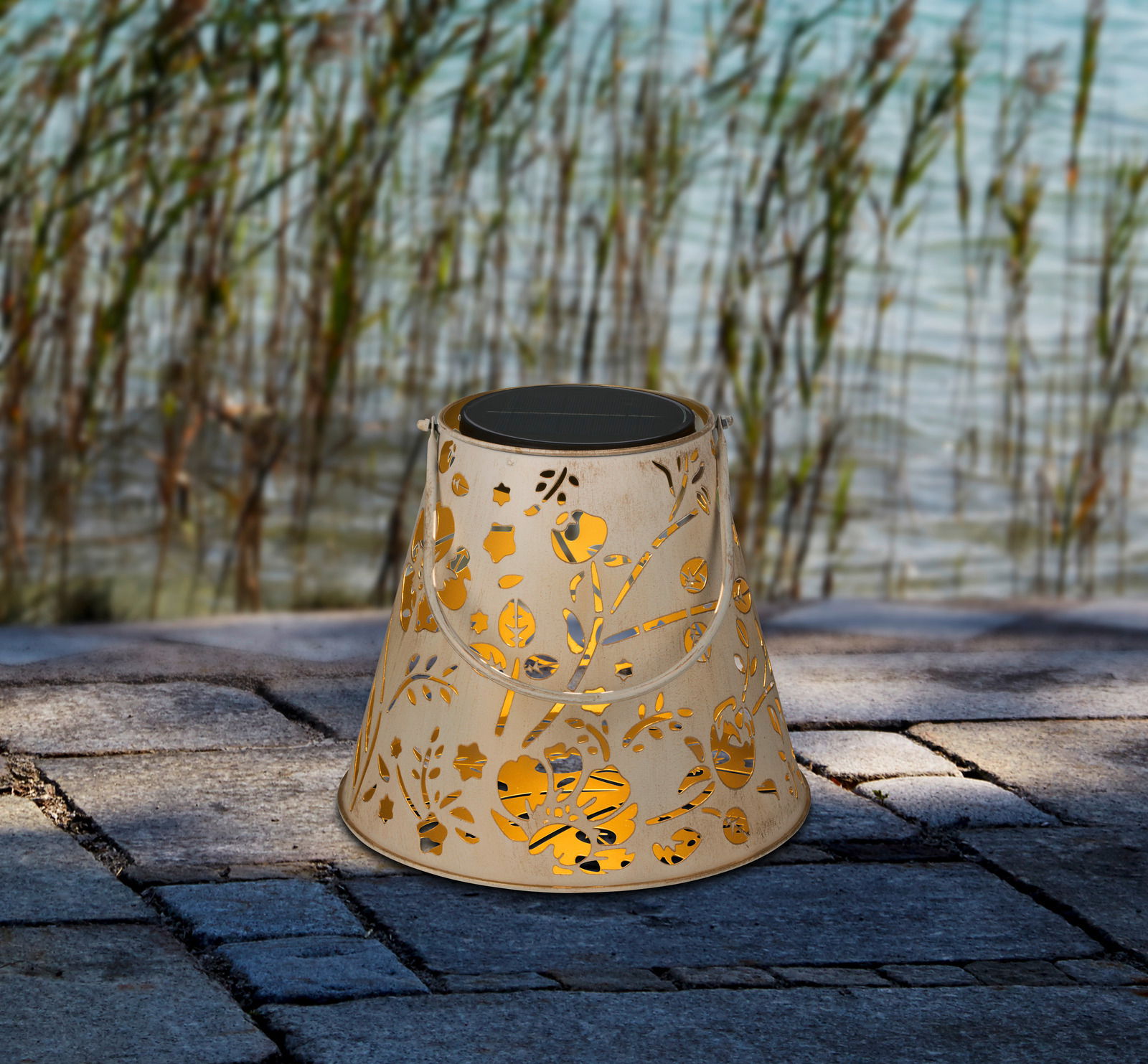 Ambia Garden LED-Solarleuchte BERN Dekorative Solarleuchte mit floralem Muster, seitliche Perspektive, auf Steinboden vor Schilf und Wasser.