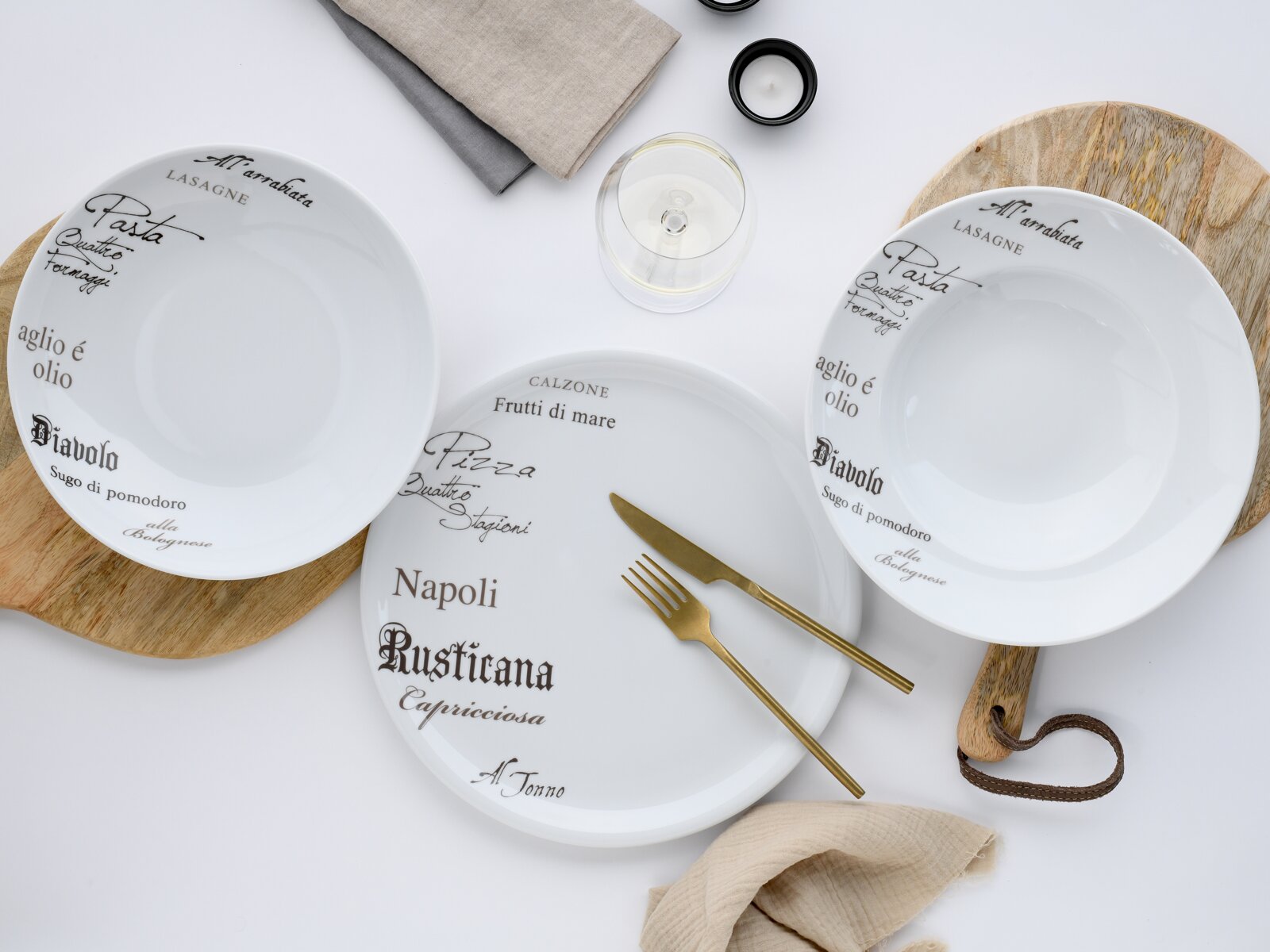 Drei weiße Porzellanteller mit italienischer Schrift in Braun und Schwarz, dekorativ auf Holzbrettchen arrangiert, mit goldfarbenem Besteck, Stoffservietten und Glas – Draufsicht von oben.