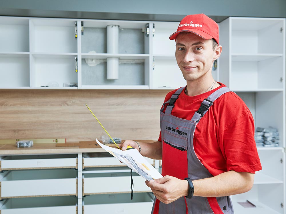 Een vakman in rode werkkleding is bezig met het monteren van een keuken; hij houdt een bouwtekening en een meetlint in zijn hand.