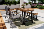 Moderner Gartentisch mit Holzplatte und Metallbeinen, umgeben von stilvollen Stühlen und einer Bank, auf einer Terrasse mit grüner Bepflanzung, aus seitlicher Perspektive fotografiert.
