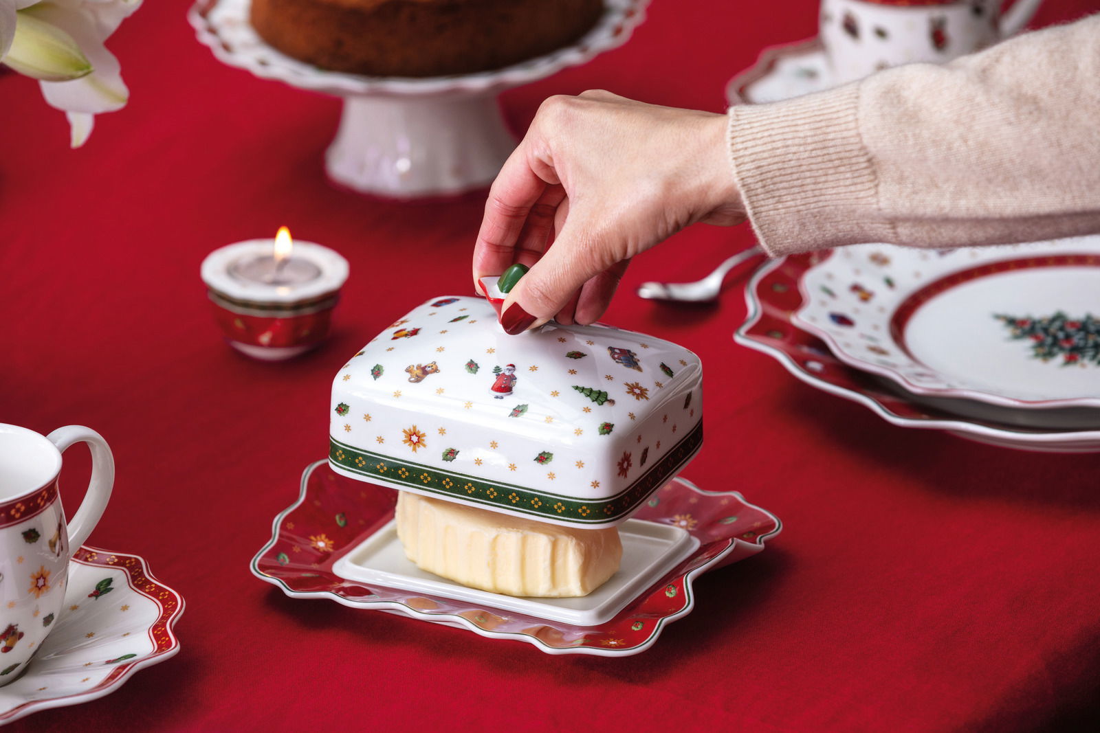 Villeroy & Boch Butterdose Hand hebt den Deckel einer festlich dekorierten Butterdose auf einem gedeckten Tisch an, seitliche Perspektive.