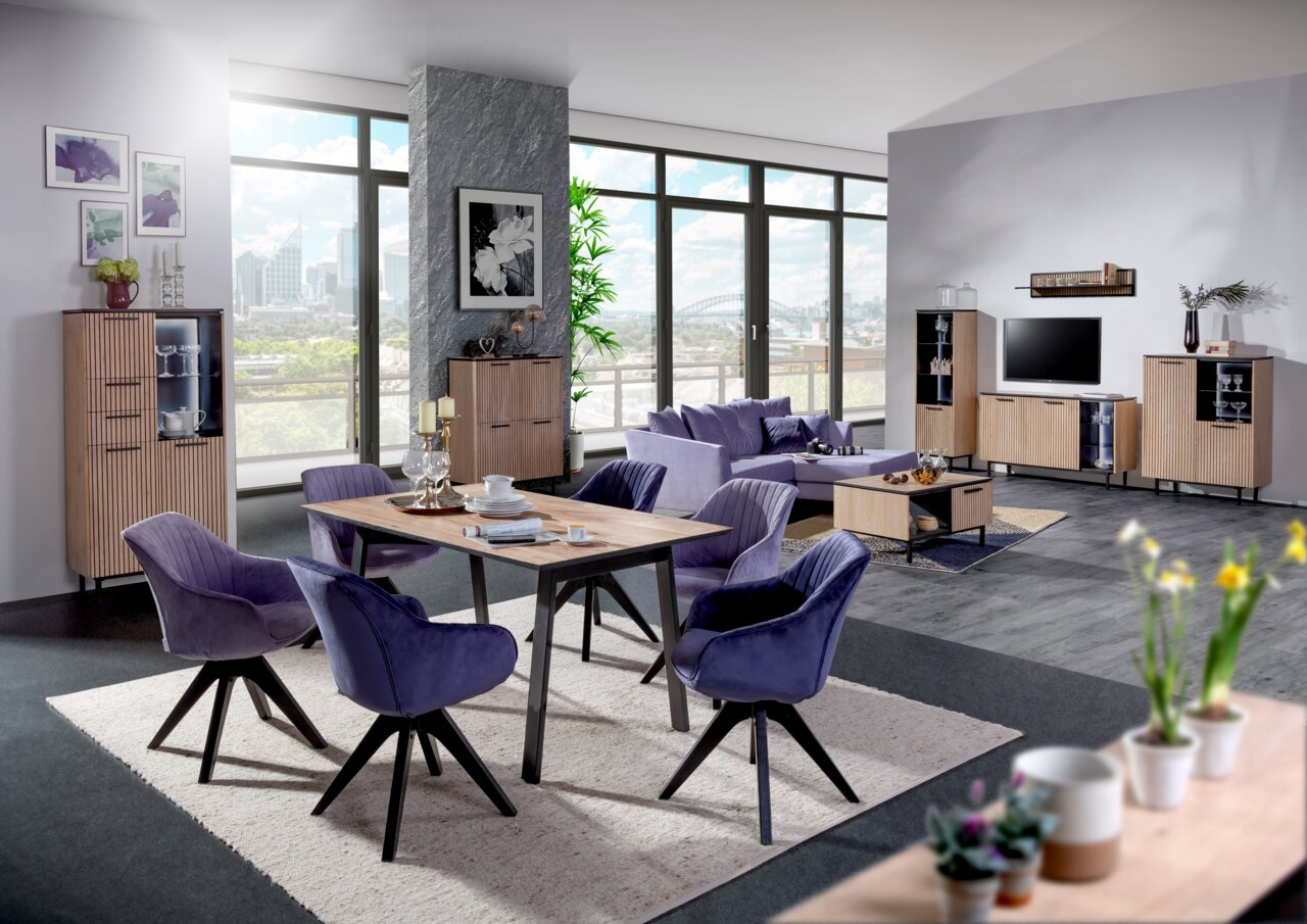 Modernes Wohnzimmer mit Esstisch und Stühlen in der Mitte, umgeben von Vitrinen, TV-Lowboard und Couchtisch in einem hellen, offenen Raum mit großen Fenstern. Perspektive von vorne.