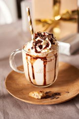 Nahaufnahme einer Glastasse mit Sahne, Schokoladensauce und einem gestreiften Strohhalm, serviert auf einem Holzteller mit Gebäckstücken.