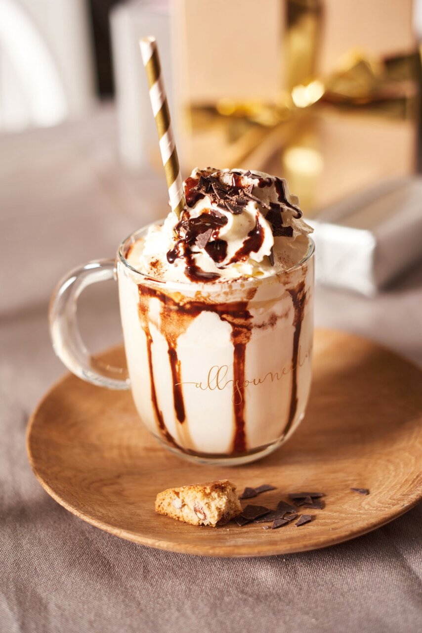 Leonardo Tasse allyouneed... Nahaufnahme einer Glastasse mit Sahne, Schokoladensauce und einem gestreiften Strohhalm, serviert auf einem Holzteller mit Gebäckstücken.