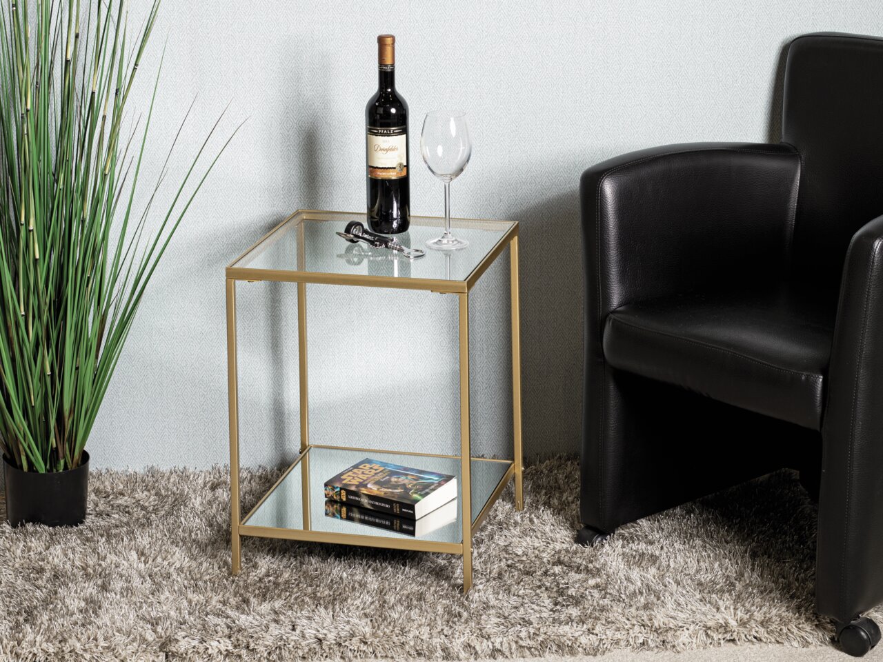 Moderner Beistelltisch mit Glasplatten und goldfarbenem Rahmen, seitliche Perspektive, dekoriert mit einer Weinflasche, einem Weinglas und Büchern, neben einem schwarzen Sessel und einer Pflanze.