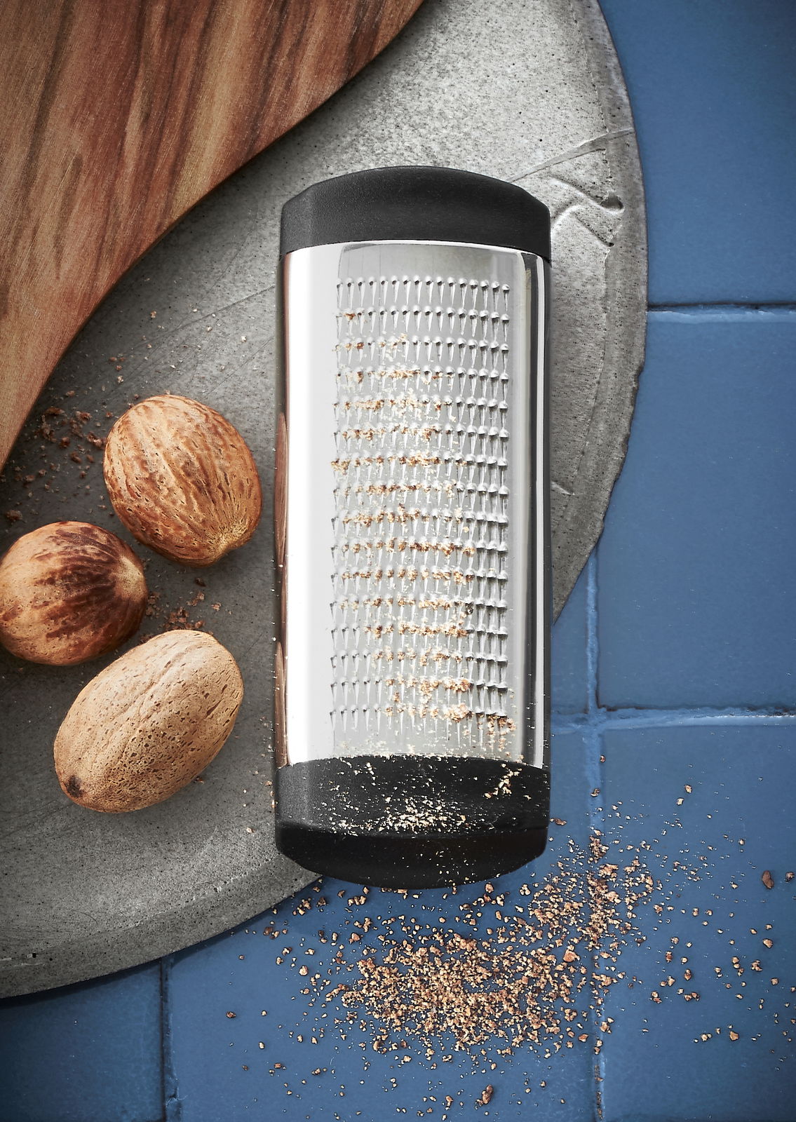 WMF Muskatreibe TOP TOOLS Muskatreibe aus Edelstahl mit schwarzem Griff, liegend auf einer blauen Fliese, umgeben von Muskatnüssen und geriebenem Muskat.