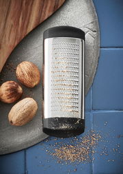 Muskatreibe aus Edelstahl mit schwarzem Griff, liegend auf einer blauen Fliese, umgeben von Muskatnüssen und geriebenem Muskat.