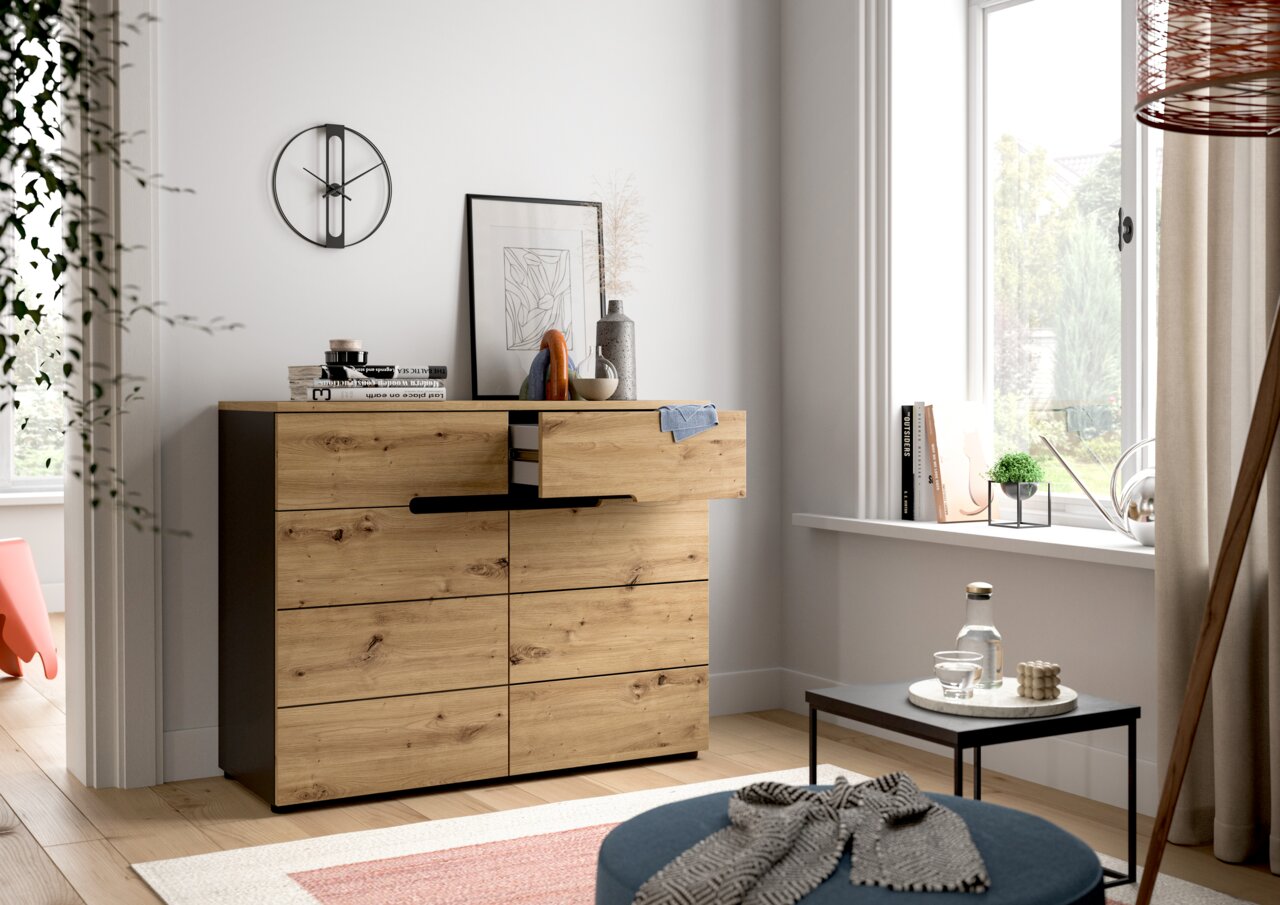 Moderne Kommode aus Holz mit Schubladen, seitliche Perspektive, dekoriert mit Büchern und Vasen, in einem hellen Raum mit Fenster.